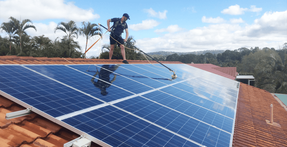 A worker cleans solar panels on a roof under a bright sky, surrounded by palm trees and hills in the background.